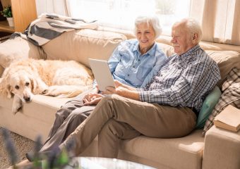 A photo of an elderly couple sat on a beige sofa with a Golden Retriever beside them.